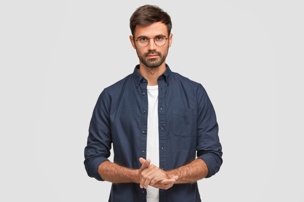 Waist Up Portrait Of Handsome Serious Unshaven Male Keeps Hands Together, Dressed In Dark Blue Shirt, Has Talk With Interlocutor, Stands Against White Background. Self Confident Man Freelancer